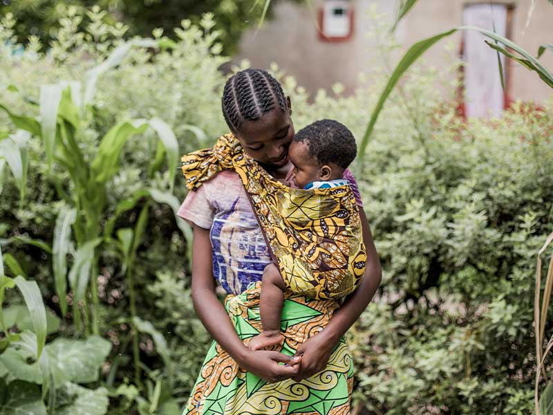 Melissa standing amongst trees/plants holding her baby son.