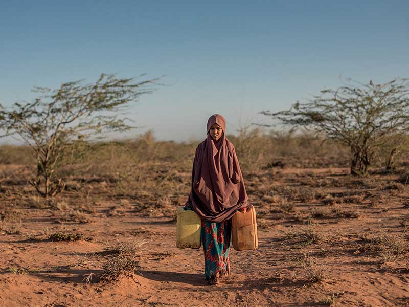 Najah carrying 2 big flasks of water across land.