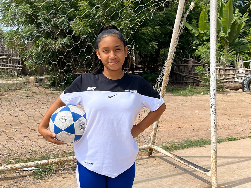 12 year old girl standing in front of goal post holding football and smiling at camera