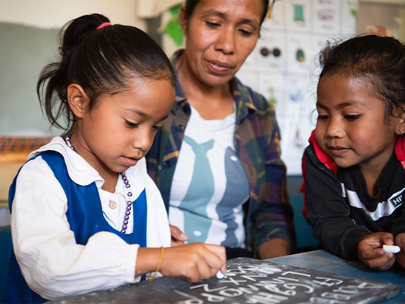 Girl practicing writing skills on chalkboard on table with another girl and her mother watching her