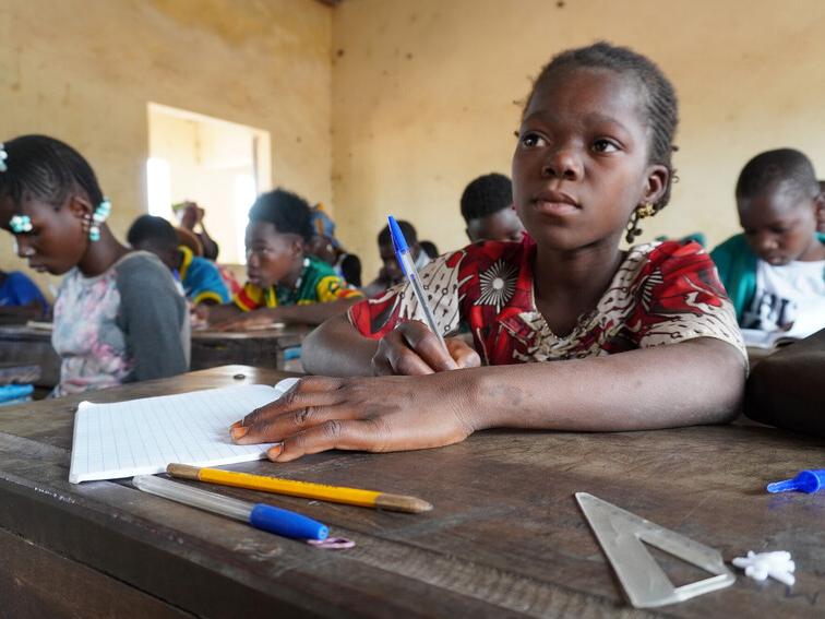 Diatou writing at school desk and looking towards the front of the classroom